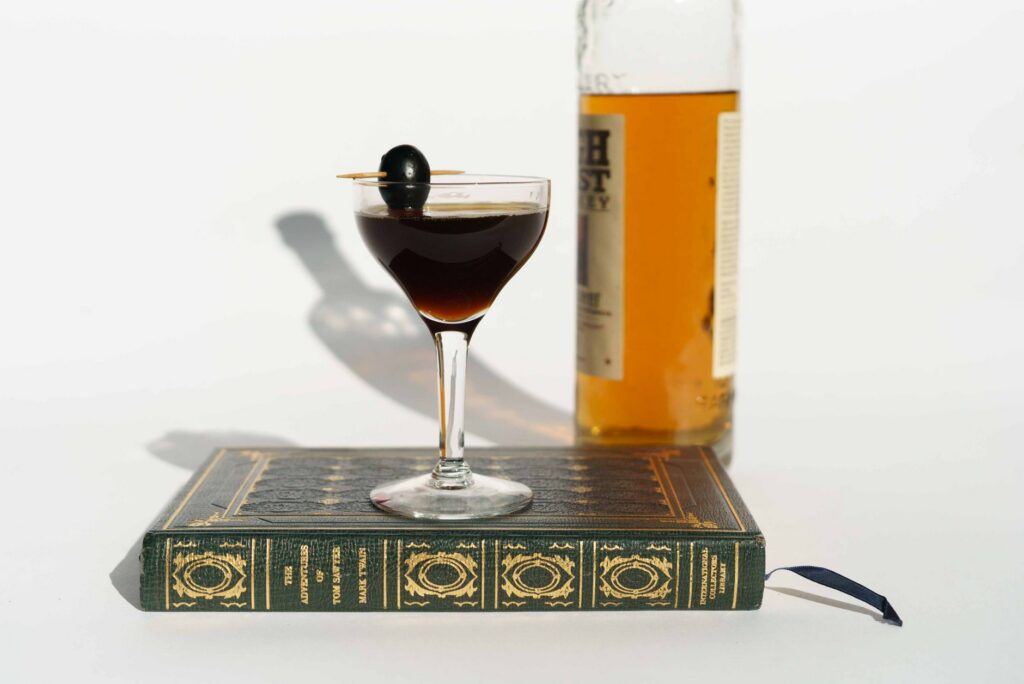Composition of glass bottle of whiskey near Manhattan cocktail served with pickled olive on stick on book
